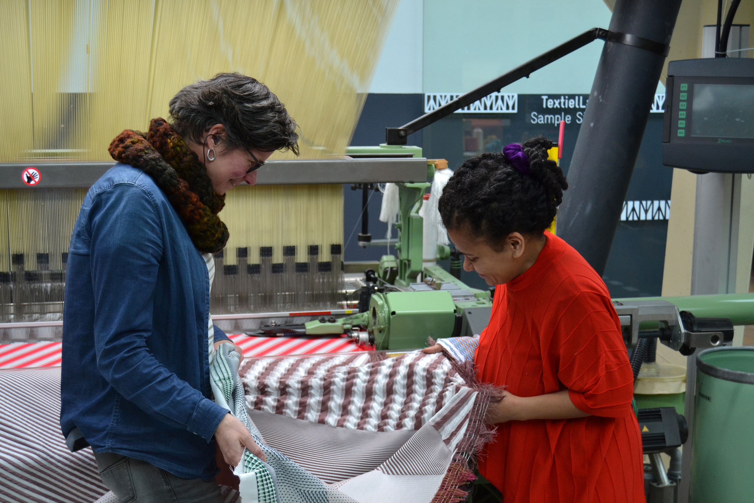 Marjan Van Oeffelt, product developer at the TextielLab and Ta'Yali Wetzel - both smiling - stand facing each other holding and examining a woven fabric that just came off the weaving machine they are stading in front of. The Jacquard harness and part of the weft rapier are visible in the background.